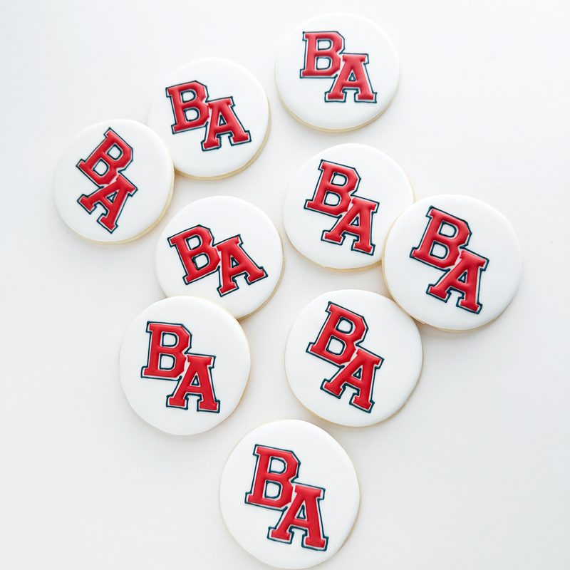 Round white cookies, each hand-iced with a red and navy collegiate monogram.