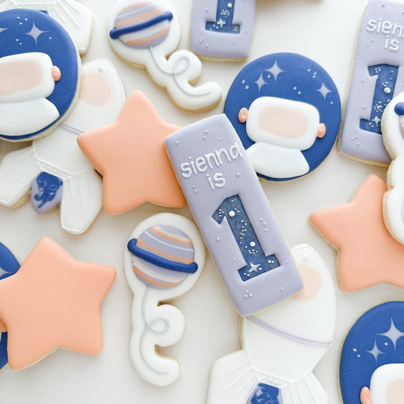 A first-birthday set in periwinkle and peach: numbered rounds, stars, rattles and a hand-lettered name.