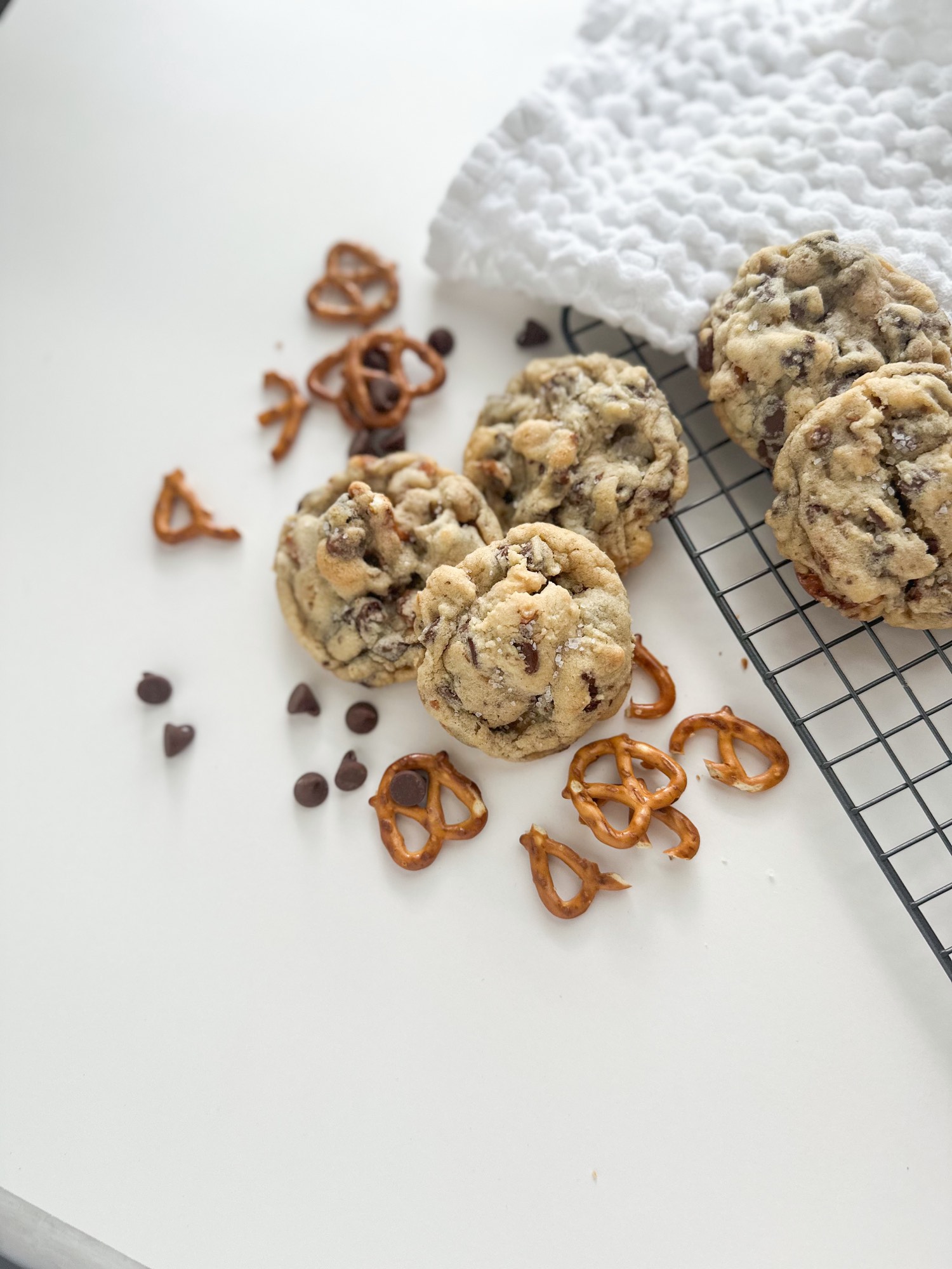 Chocolate chip pretzel cookies on a cooling rack beside a white waffle towel, pretzels scattered across the table.
