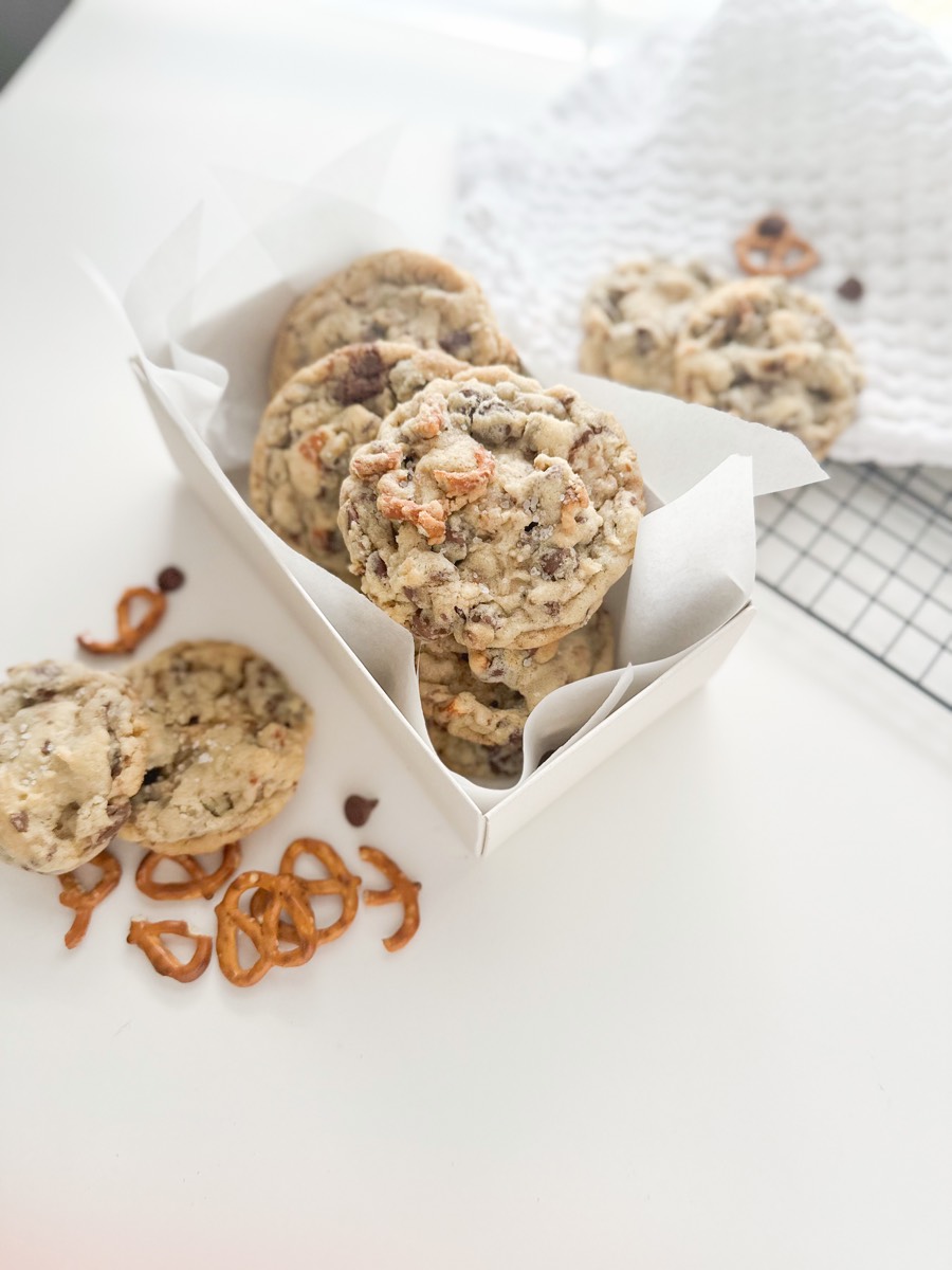 Loaded cookies with pretzels and chocolate chunks piled in a white box, more resting beside it.