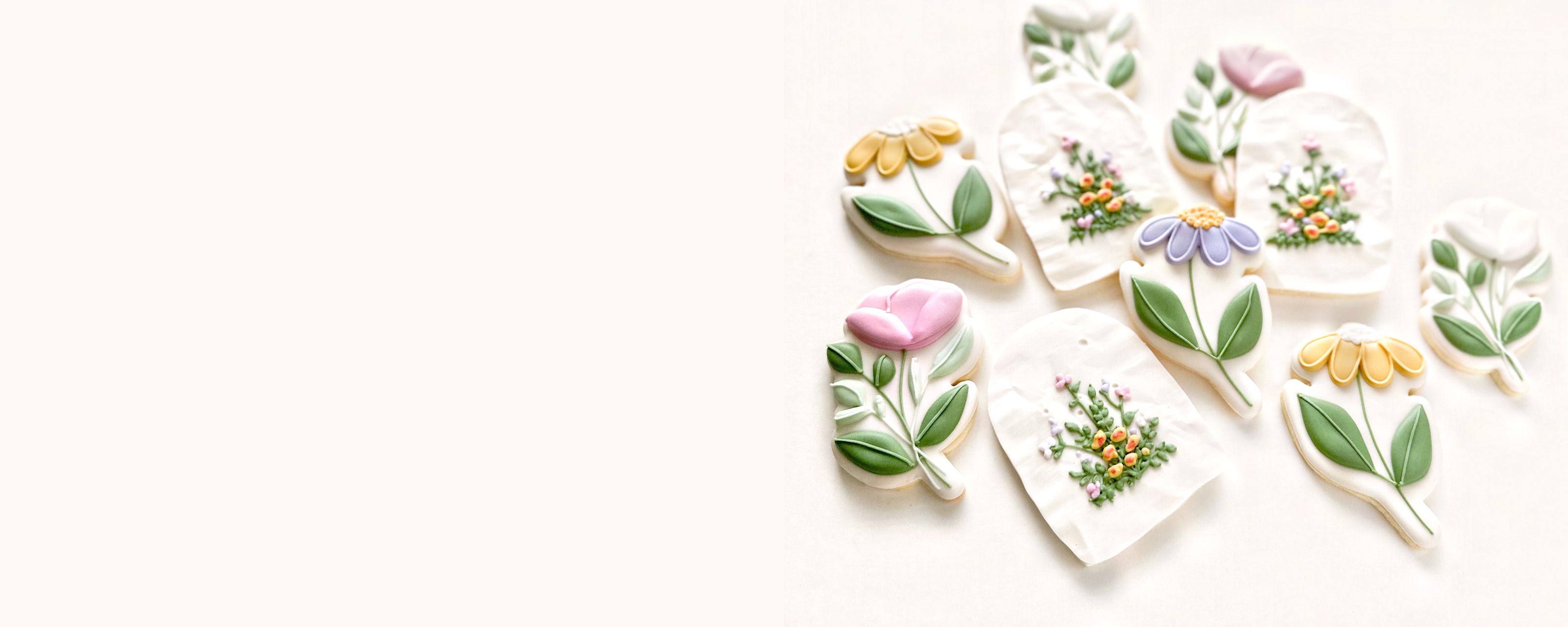 Hand-iced cookies shaped as tulips, daisies and pressed-flower plaques in pale green, lilac, pink and yellow, arranged on a pale grey surface.
